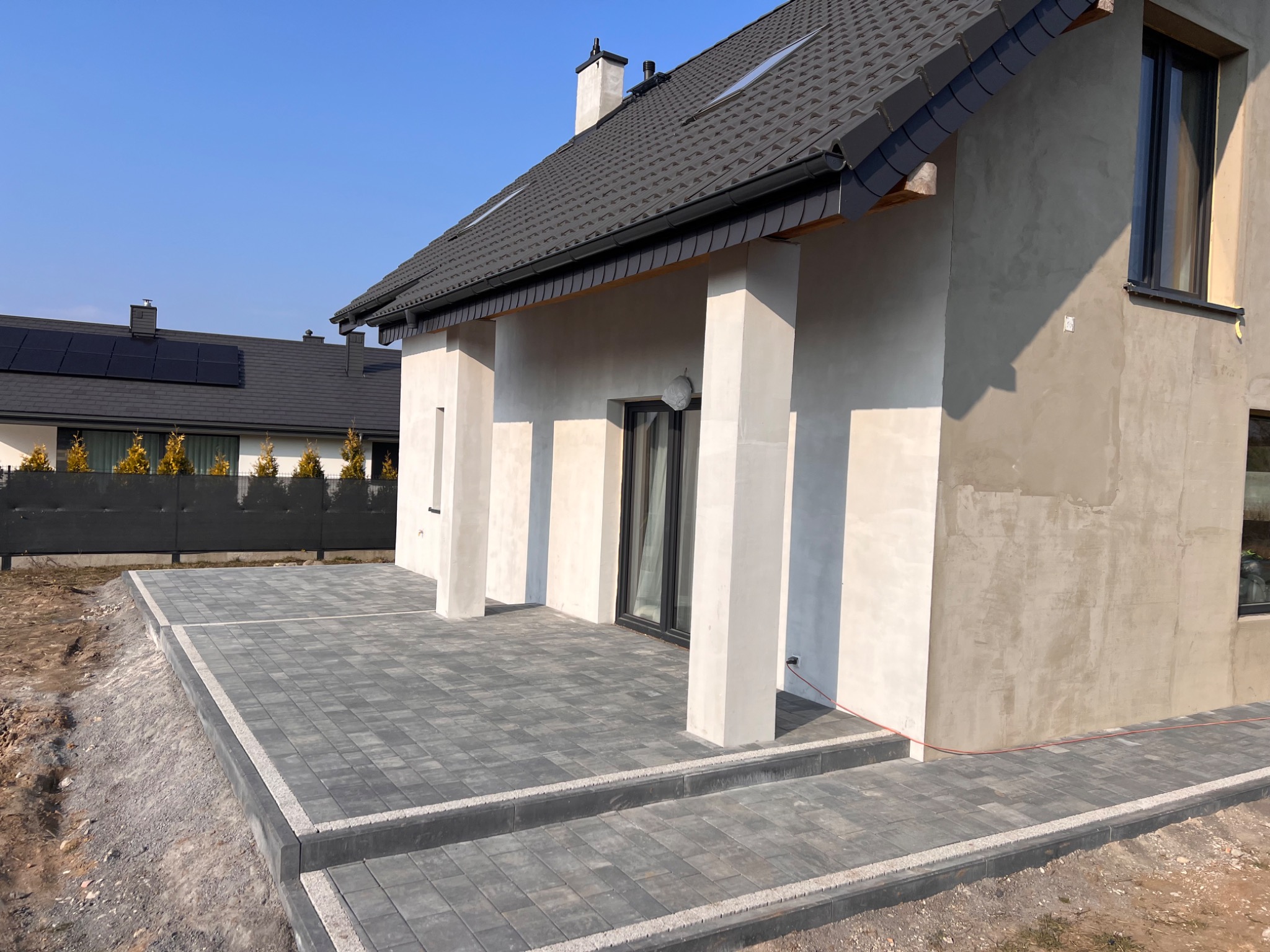 Nowoczesny dom z szarą kostką brukową na tarasie i schodach wejściowych, widok na elewację w trakcie wykańczania i fragment dachu z czarną dachówką.