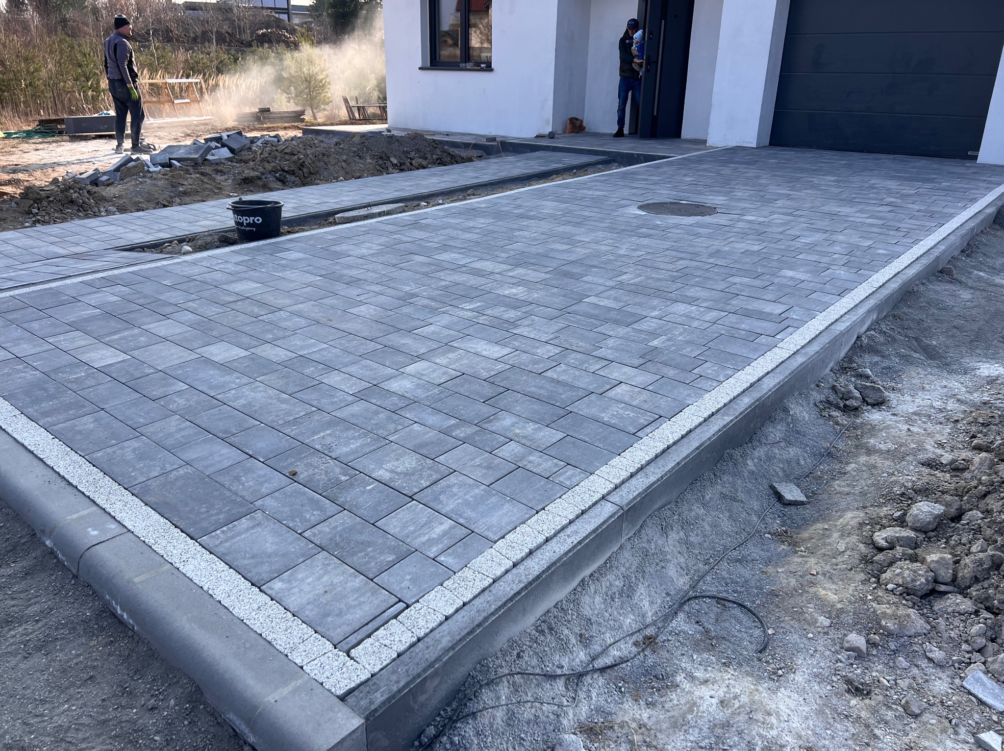 Nowo ułożony podjazd z szarej kostki brukowej przed nowoczesnym domem, obrzeża wykończone jasnym kamieniem, w tle pracownik przy rozplantowywaniu ziemi.