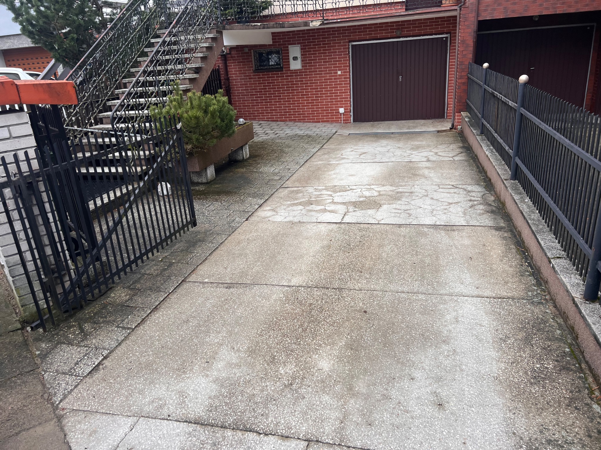 Betonowy podjazd do garażu z widocznymi pęknięciami i śladami użytkowania. Po bokach metalowe ogrodzenie, schody na piętro i dekoracyjna zieleń w donicy.