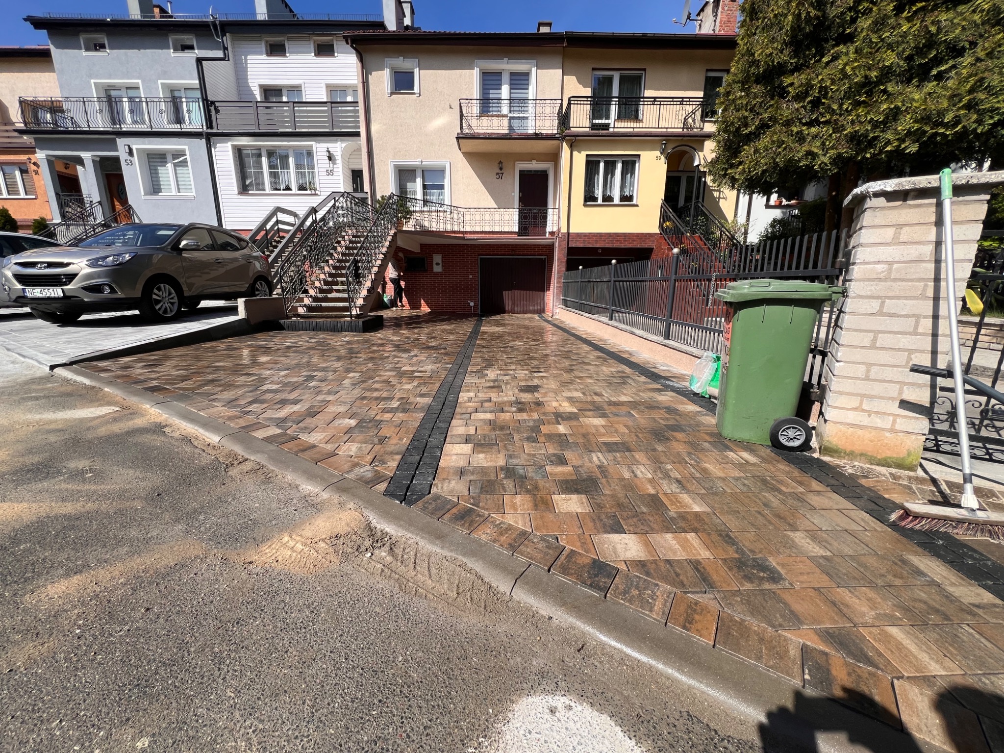 Ułożona kostka brukowa w kolorach ziemi tworzy wzorzysty podjazd do garażu. Widoczne domy w tle i samochód. Na pierwszym planie krawężnik oddzielający kostkę od ulicy.