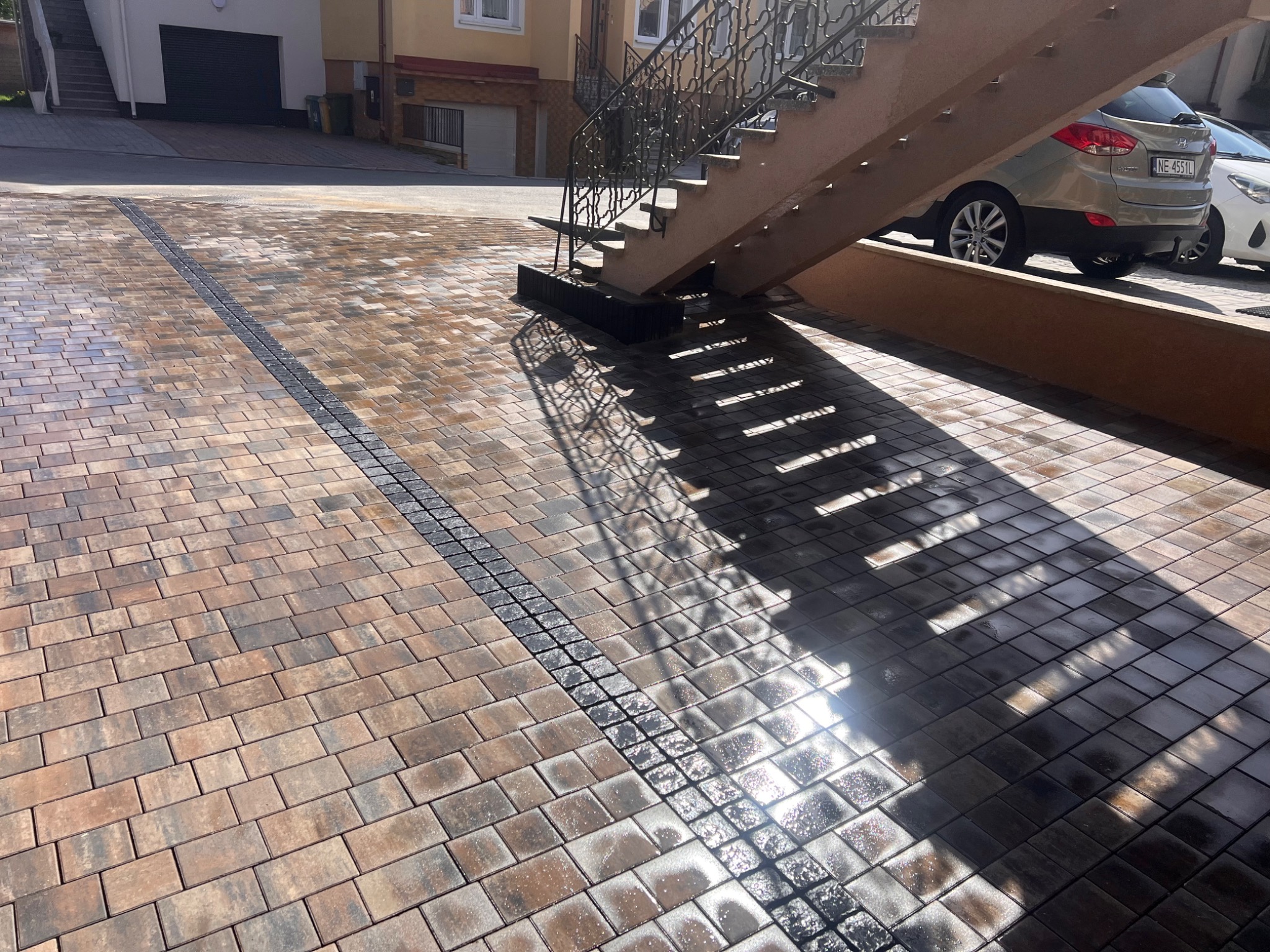 Ułożona kostka brukowa w Lubawie z odpływem liniowym. Widoczne schody i cień rzucany przez barierki. Kostka w odcieniach brązu i szarości, lekko mokra.