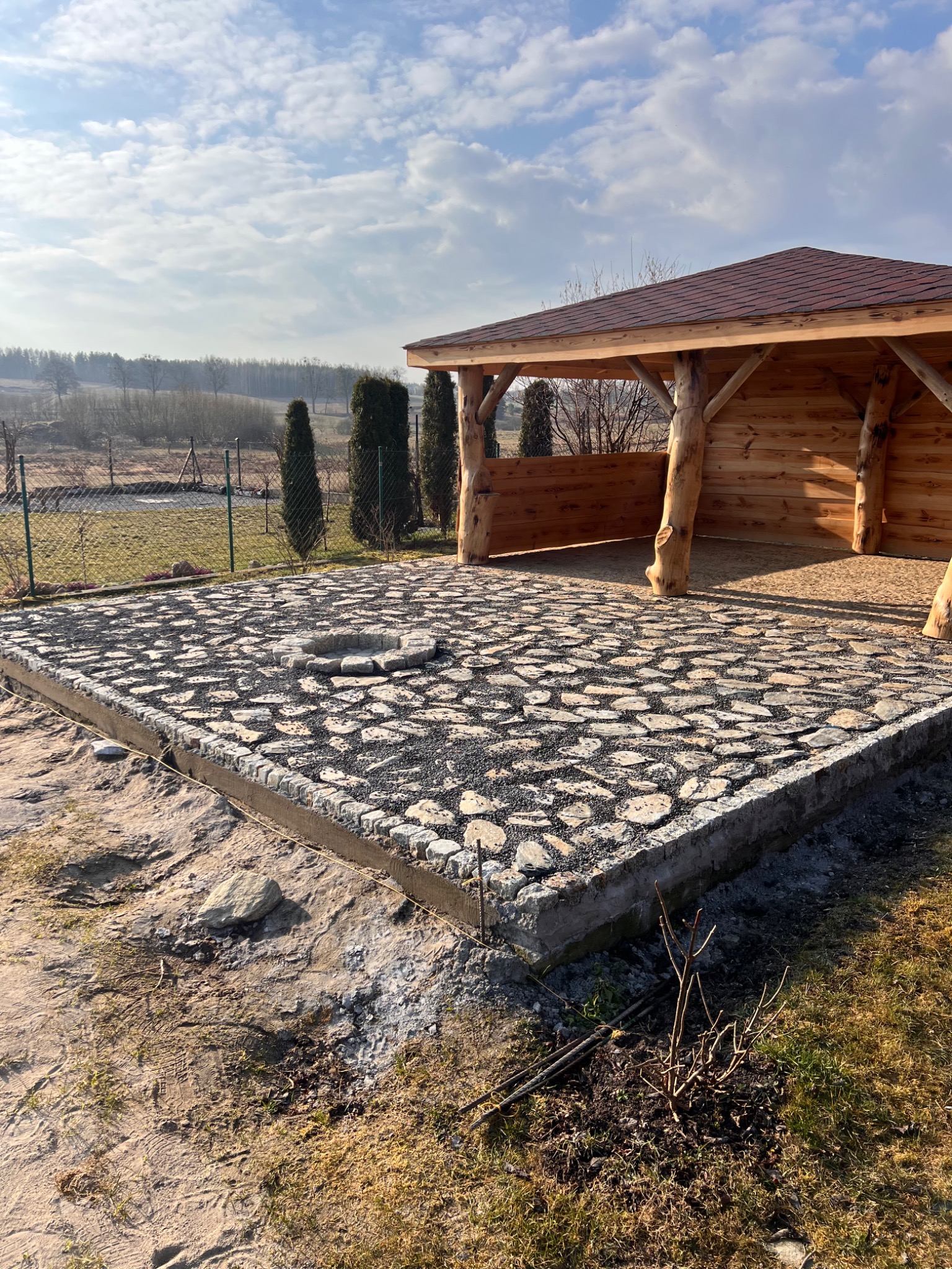 Granitowa podłoga tarasu z paleniskiem w rustykalnej altanie, otoczonej zielenią. Widok na pagórkowaty krajobraz pod błękitnym niebem z chmurami.