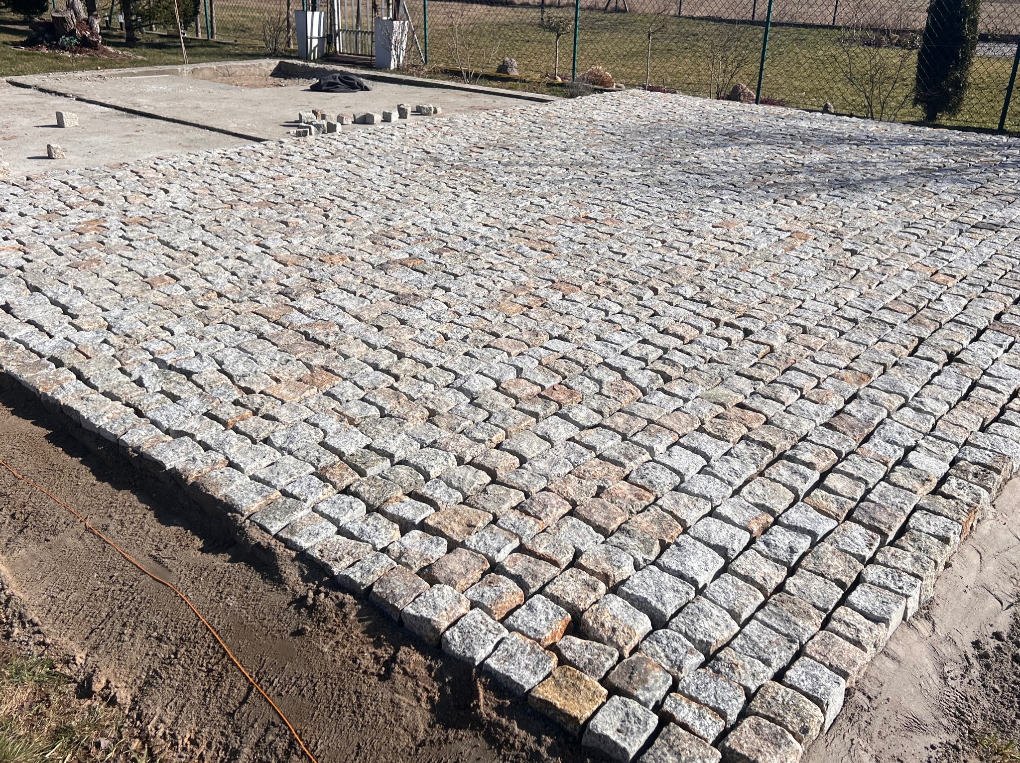 Ułożona kostka granitowa w regularny wzór, widoczne fragmenty terenu przed i za. Częściowo ułożona nawierzchnia z jasnoszarej kostki.