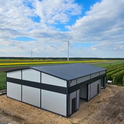 STEELGUYS SPÓŁKA Z OGRANICZONĄ ODPOWIEDZIALNOŚCIĄ - Gotowa hala stalowa z panelami w kolorze szarym i czarnym, widok z góry na tle pól uprawnych, turbin wiatrowych i podnośnika koszowego z pracownikiem.