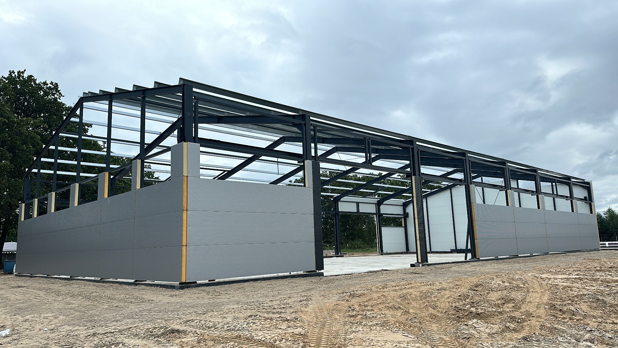 Konstrukcja stalowa hali w trakcie budowy, widoczne szkielet i częściowo zamontowane szare panele ścienne z blachy trapezowej, teren budowy z piaskiem.