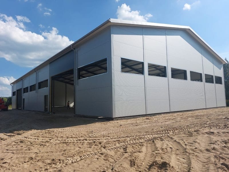 Nowoczesna hala przemysłowa z szarej blachy trapezowej, widoczna konstrukcja stalowa przez otwory okienne, teren wokół wyrównany, w tle błękitne niebo z chmurami.