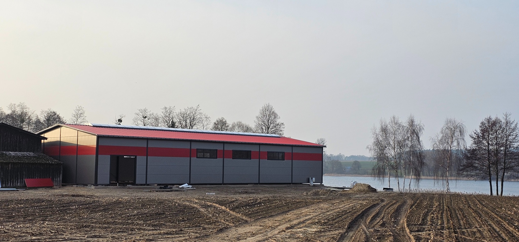 Nowoczesna hala stalowa w odcieniach szarości z czerwonym akcentem, położona na skraju zaoranego pola w pobliżu jeziora, widoczna z zewnątrz w pochmurny dzień.