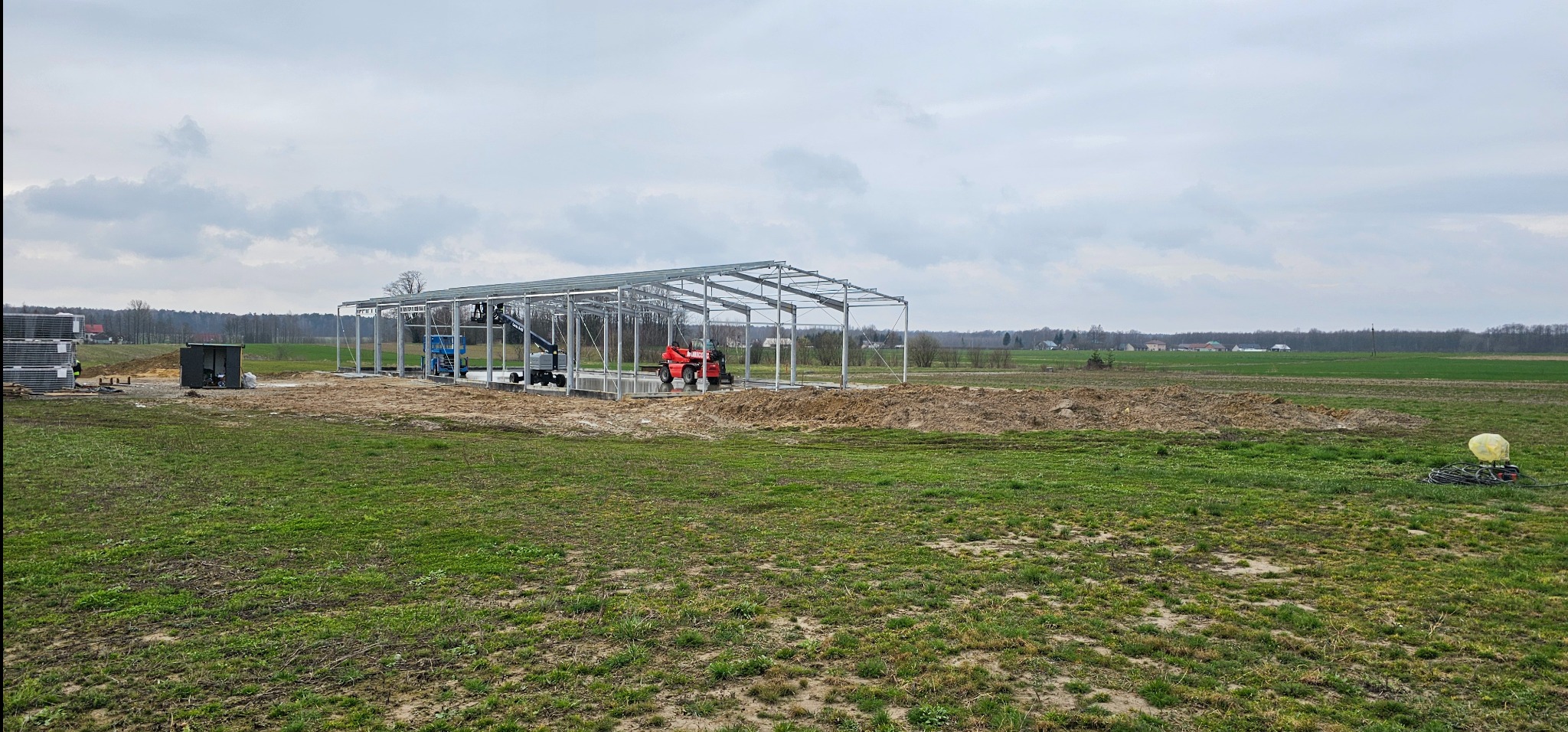 Montaż konstrukcji stalowej hali na terenie gospodarstwa rolnego, widoczne podnośniki i materiały budowlane, pochmurne niebo.