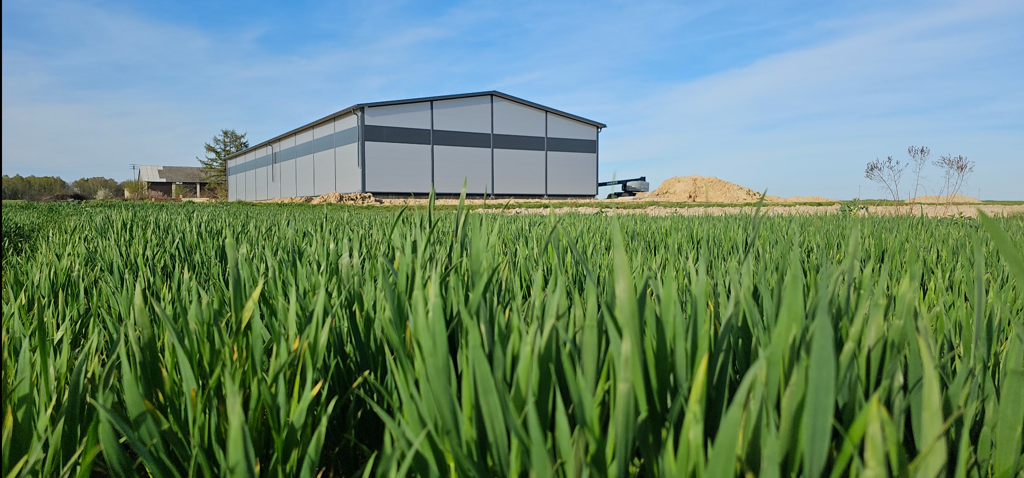 Nowoczesna hala magazynowa z szarej blachy trapezowej z ciemnoszarym pasem w górnej części, widziana z poziomu niskiej trawy na polu, w tle kopiec ziemi i podnośnik koszowy.