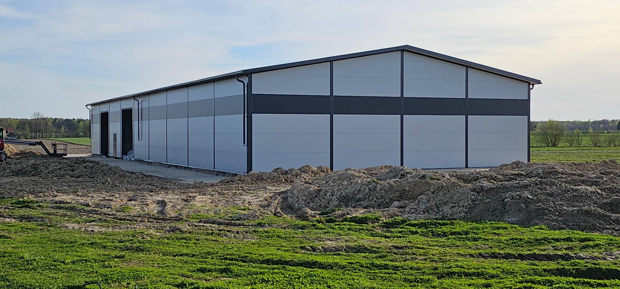 Nowoczesna hala magazynowa z panelami w odcieniach szarości, widoczna z zewnątrz, z niezagospodarowanym terenem wokół i sprzętem budowlanym w tle.