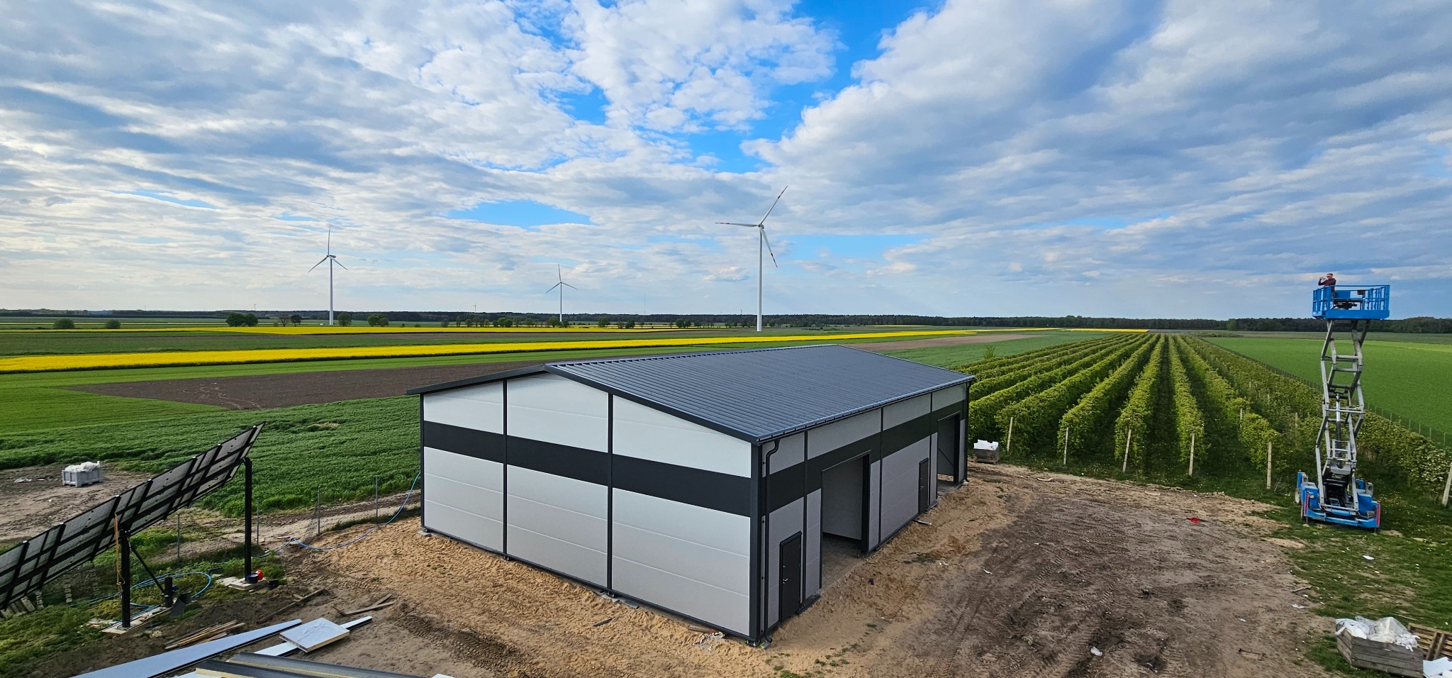 Gotowa hala stalowa z panelami w kolorze szarym i czarnym, widok z góry na tle pól uprawnych, turbin wiatrowych i podnośnika koszowego z pracownikiem.