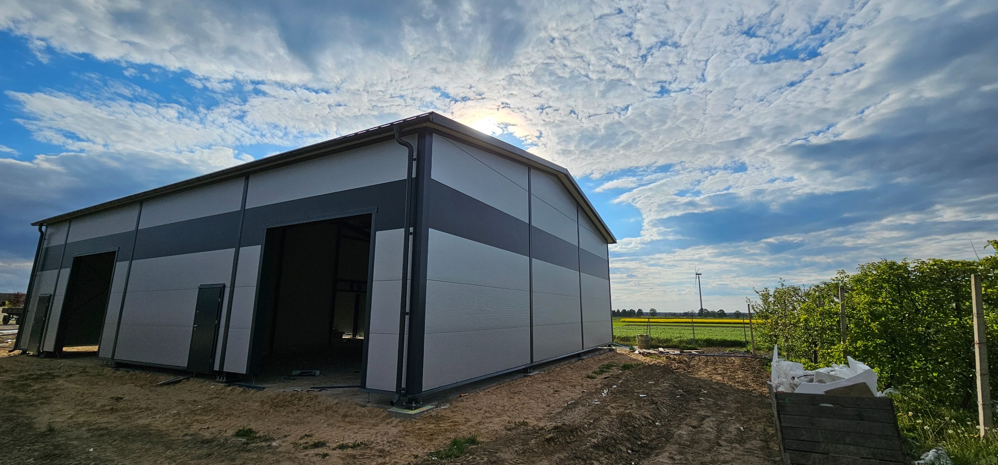 Nowo wybudowana hala z blachy trapezowej w odcieniach szarości, z widocznym otwartym wjazdem i drzwiami, na tle błękitnego nieba z chmurami i zielenią w tle.