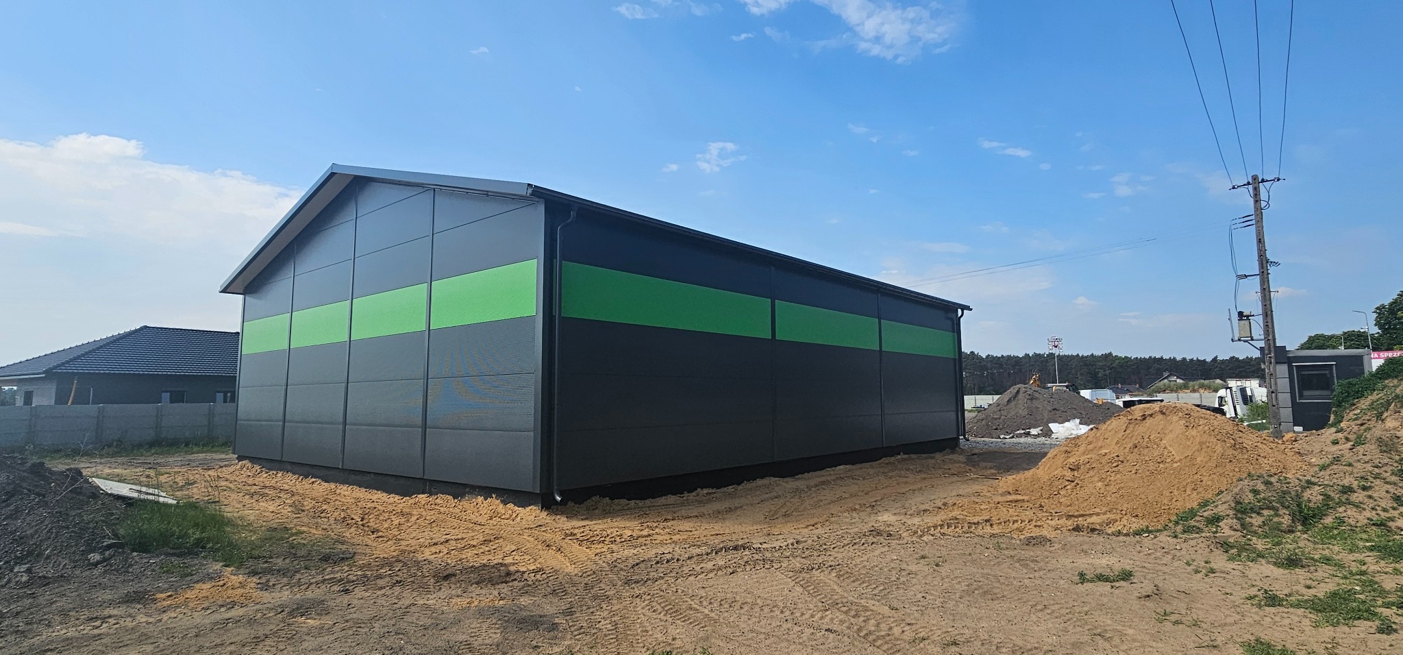 Nowoczesna hala magazynowa z szarymi panelami i poziomym zielonym pasem, widok z zewnątrz w słoneczny dzień, teren wokół w trakcie prac ziemnych.