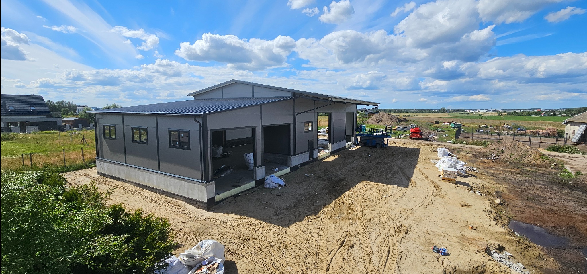 Wznoszona hala stalowa z szarej blachy trapezowej, widok zewnętrzny z boku, teren budowy z piaskiem i sprzętem budowlanym w tle, błękitne niebo z chmurami.