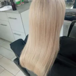 Długie, proste włosy w odcieniu chłodnego blondu, widoczne od tyłu na czarnym fotelu w salonie, z refleksami światła na końcówkach.