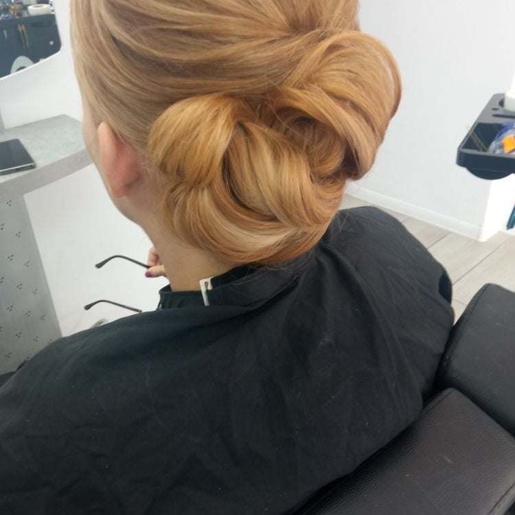 Upięcie włosów w kok z tyłu głowy, włosy w odcieniach blondu z refleksami, widoczny czarny fartuch ochronny i fragment okularów.