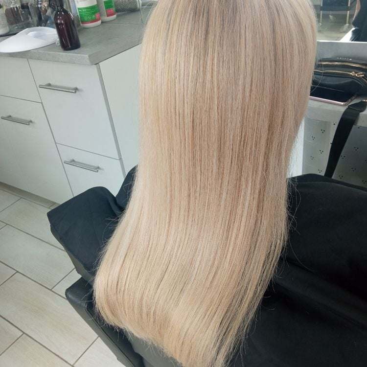 Długie, proste włosy w odcieniu chłodnego blondu, widoczne od tyłu na czarnym fotelu w salonie, z refleksami światła na końcówkach.