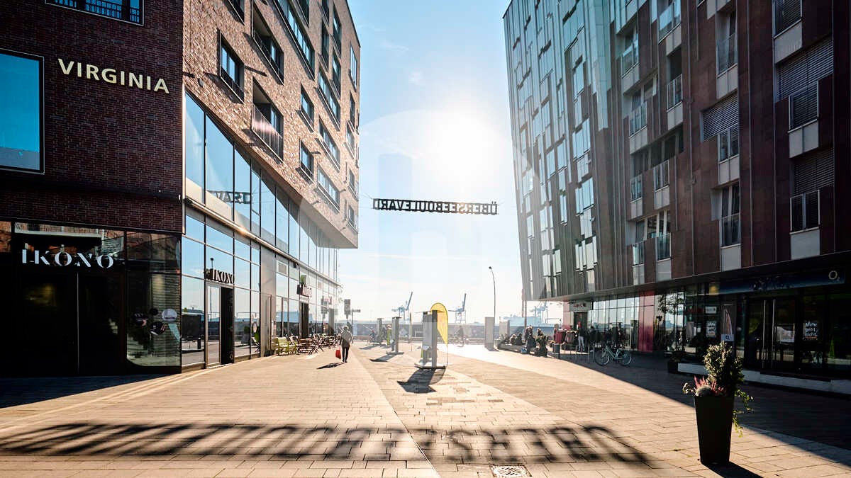 Ulica miejska z nowoczesną architekturą, widoczne budynki z dużymi oknami, chodnik z kostki brukowej i dekoracyjne napisy nad ulicą.