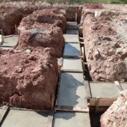 Wykopane fundamenty z drewnianym szalunkiem wypełnione świeżym betonem, otoczone czerwoną ziemią, w tle widoczne drzewa i żółte kwiaty.