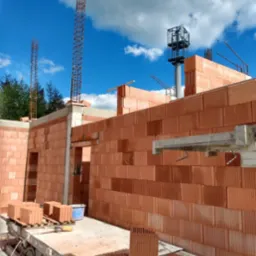 Wznoszenie ścian domu z czerwonej ceramiki poryzowanej, widoczne zbrojenia słupów, elementy rusztowania i paleta z bloczkami, w tle błękitne niebo z chmurami.