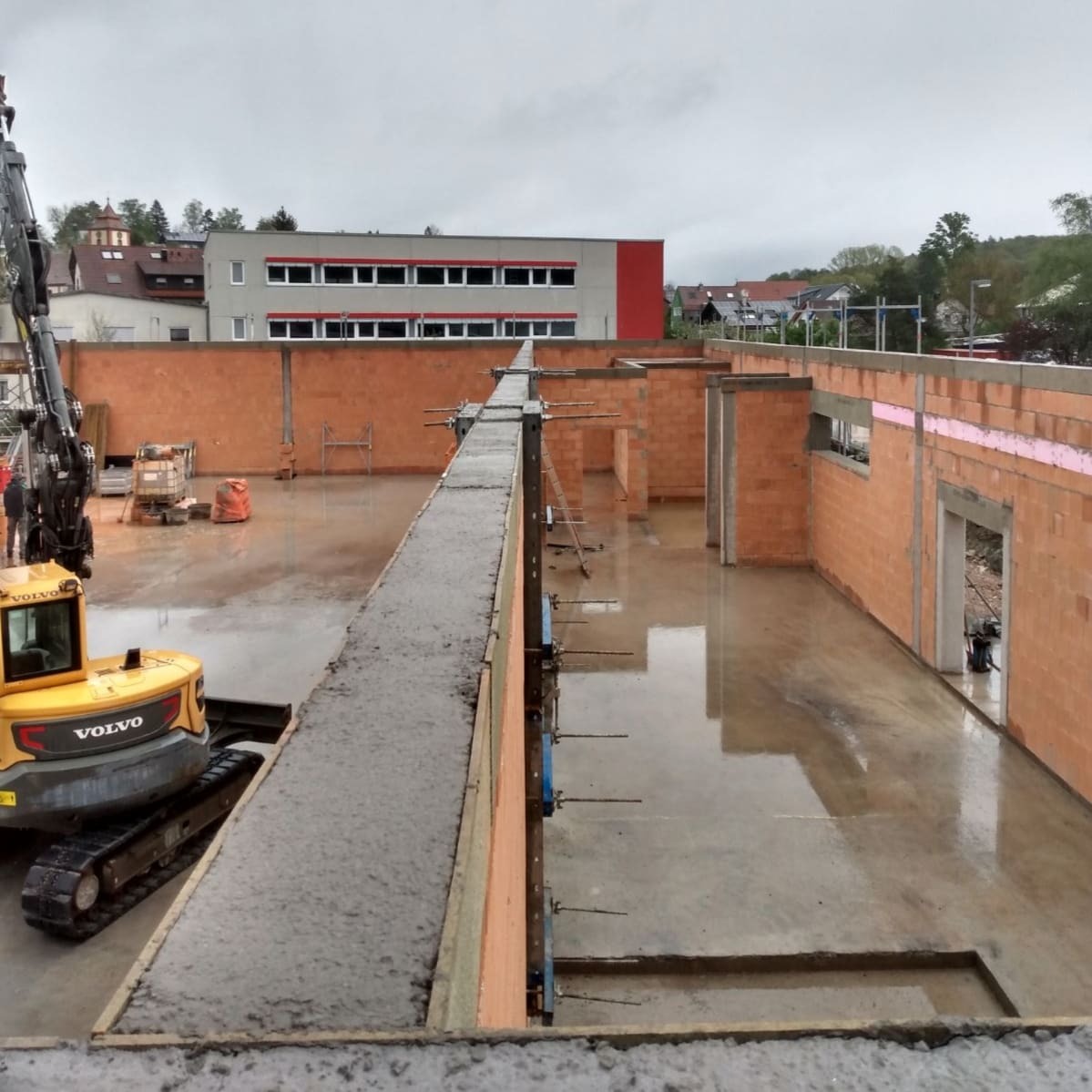Budowa domu w trakcie wznoszenia ścian z cegły ceramicznej, widoczny zalany betonem wieniec, żółta koparka Volvo oraz mokra posadzka.