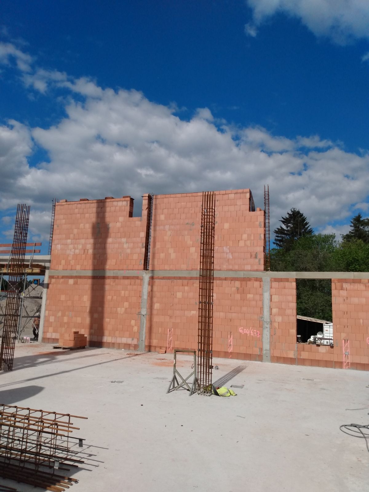 Wznoszone ściany domu z czerwonej cegły ceramicznej, zbrojenie pionowe wystające ponad mur, betonowe nadproża, błękitne niebo z chmurami w tle.