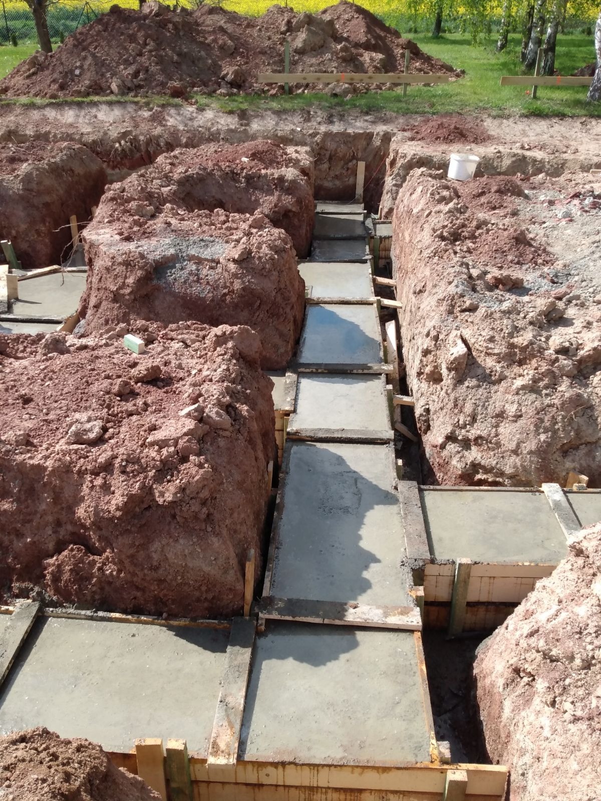 Wykopane fundamenty z drewnianym szalunkiem wypełnione świeżym betonem, otoczone czerwoną ziemią, w tle widoczne drzewa i żółte kwiaty.