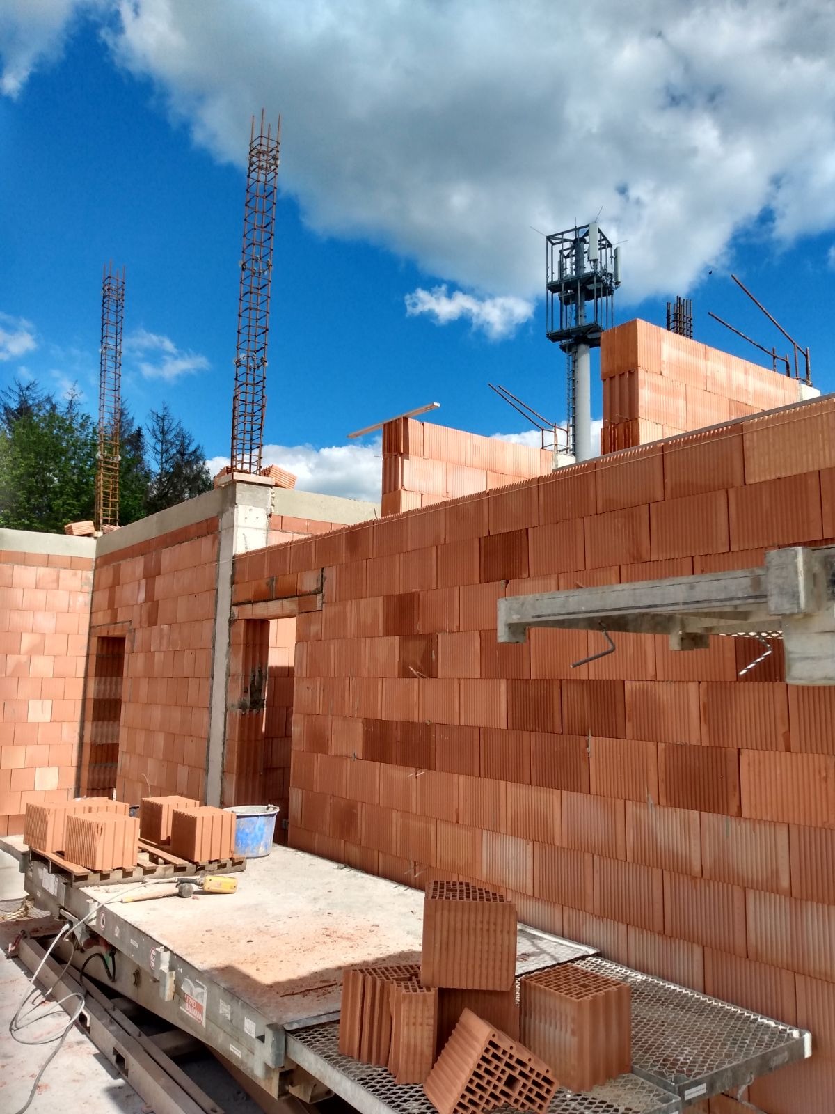 Wznoszenie ścian domu z czerwonej ceramiki poryzowanej, widoczne zbrojenia słupów, elementy rusztowania i paleta z bloczkami, w tle błękitne niebo z chmurami.