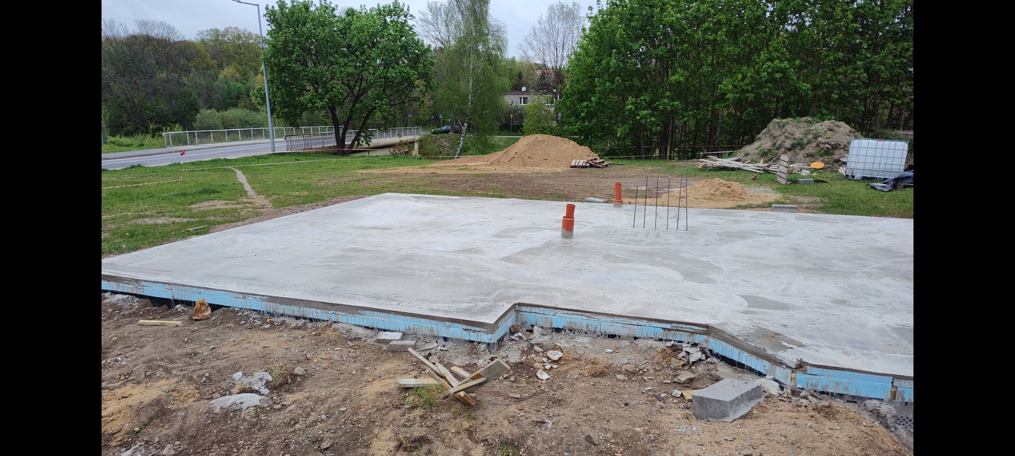 Wylana płyta fundamentowa z wystającymi rurami kanalizacyjnymi i prętami zbrojeniowymi, zabezpieczona niebieskim styropianem, na tle zieleni i drogi.