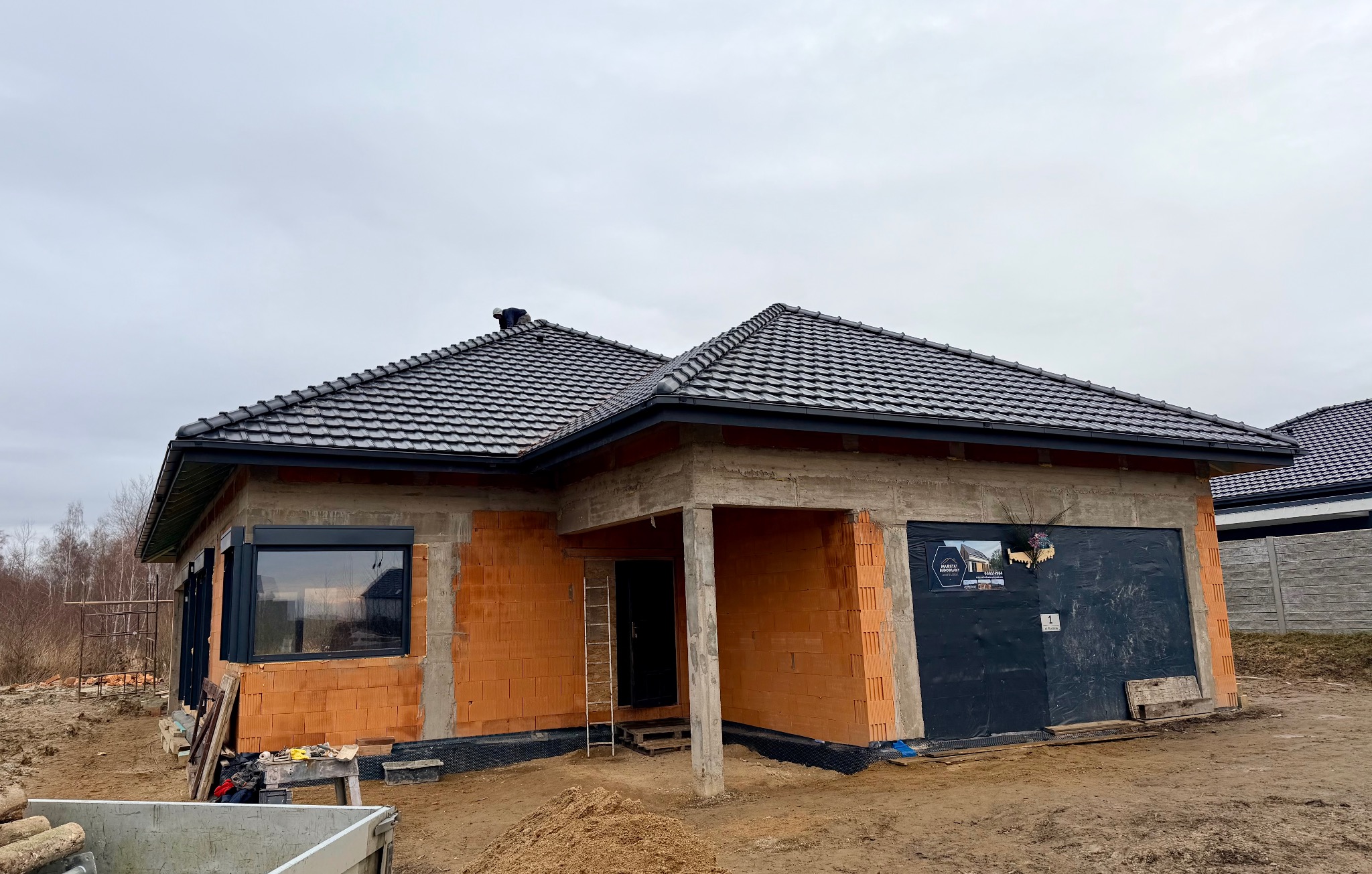 Niedokończony dom z czerwonej cegły z czarnym dachem i oknami. Widoczne rusztowania i drabina. Garaż zabezpieczony czarną folią z reklamą. Szary dzień.