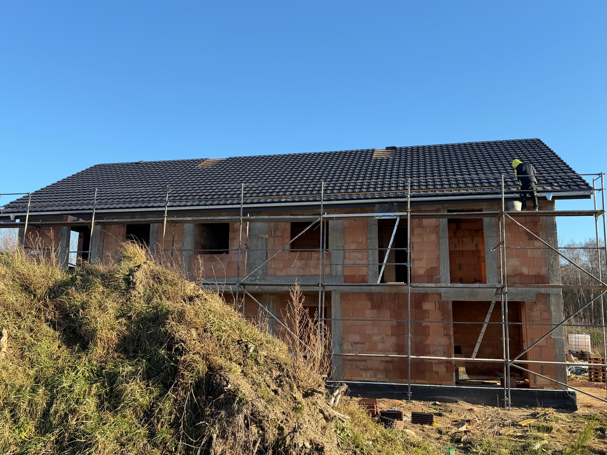 Dom w trakcie budowy z czerwonej cegły, z czarnym dachem i rusztowaniem. Na dachu pracuje osoba w żółtej czapce. W tle błękitne niebo.