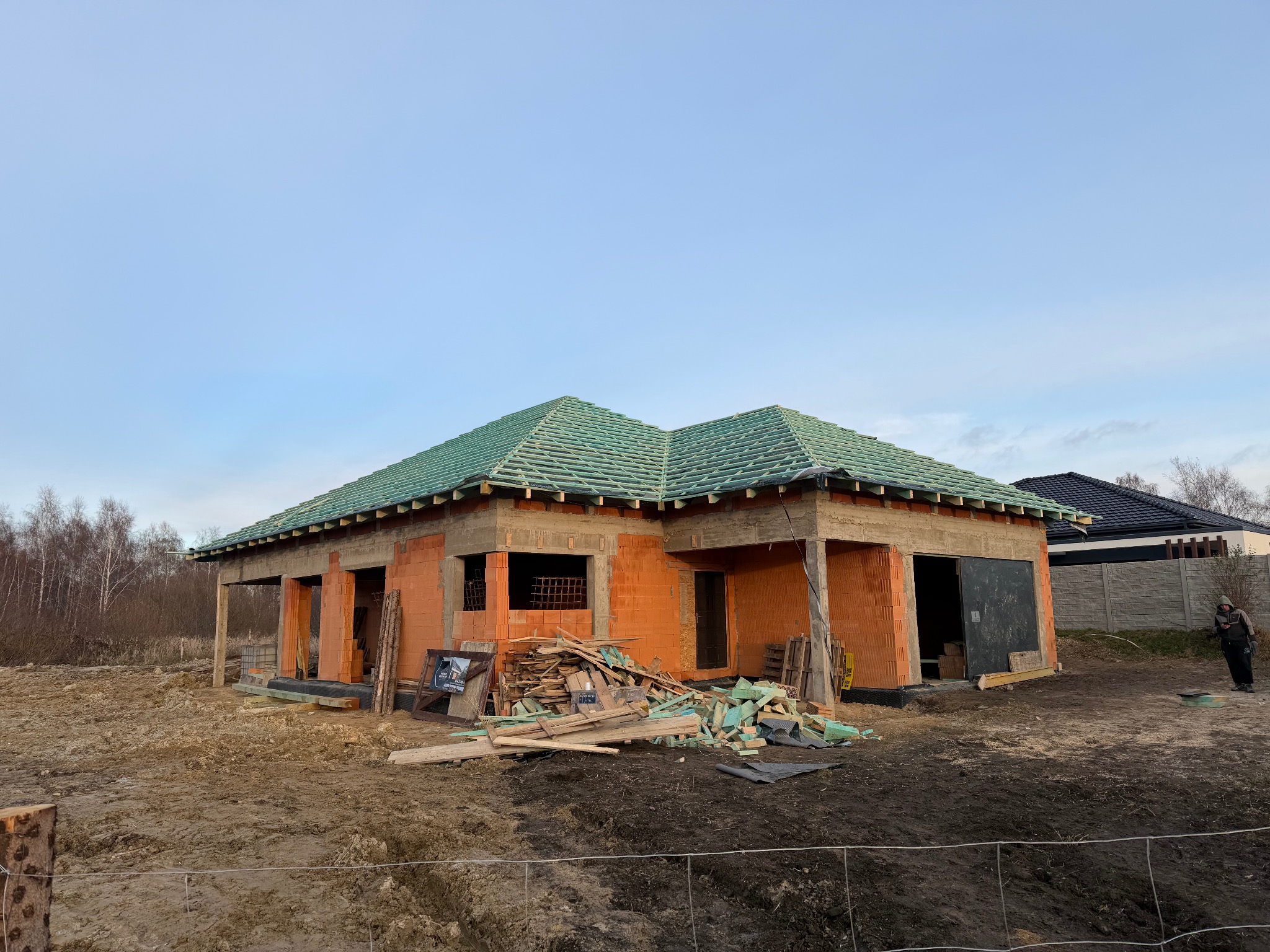 Niedokończony dom z czerwonej cegły z zielonym dachem, widok z zewnątrz, teren budowy z gruzem i błotem, w tle drzewa i niebo.