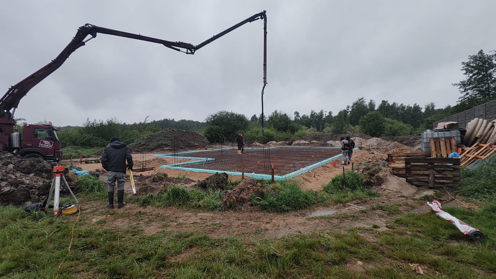 Wylewka fundamentów domu jednorodzinnego z użyciem pompy do betonu. Zbrojenie, izolacja termiczna i geodezyjne pomiary w toku. Widok na plac budowy w pochmurny dzień.