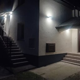 Sylwester Dyguła - Oświetlony budynek z zewnętrznymi schodami i wejściem, nocne ujęcie z niskim kątem, widoczne detale elewacji i oświetlenia.