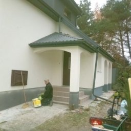 Sylwester Dyguła - Pracownik z wiadrem farby klęczy przed wejściem do domu z pomalowaną elewacją. Widoczne narzędzia i fragmenty rusztowania. Świeżo pomalowana elewacja w jasnym odcieniu.