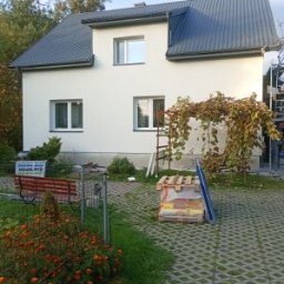 Sylwester Dyguła - Biały dom z szarą blachodachówką po malowaniu elewacji, widok z ogrodu z kwiatami i ławką. Na ścianie pnącze, obok rusztowanie. Widoczne materiały budowlane.