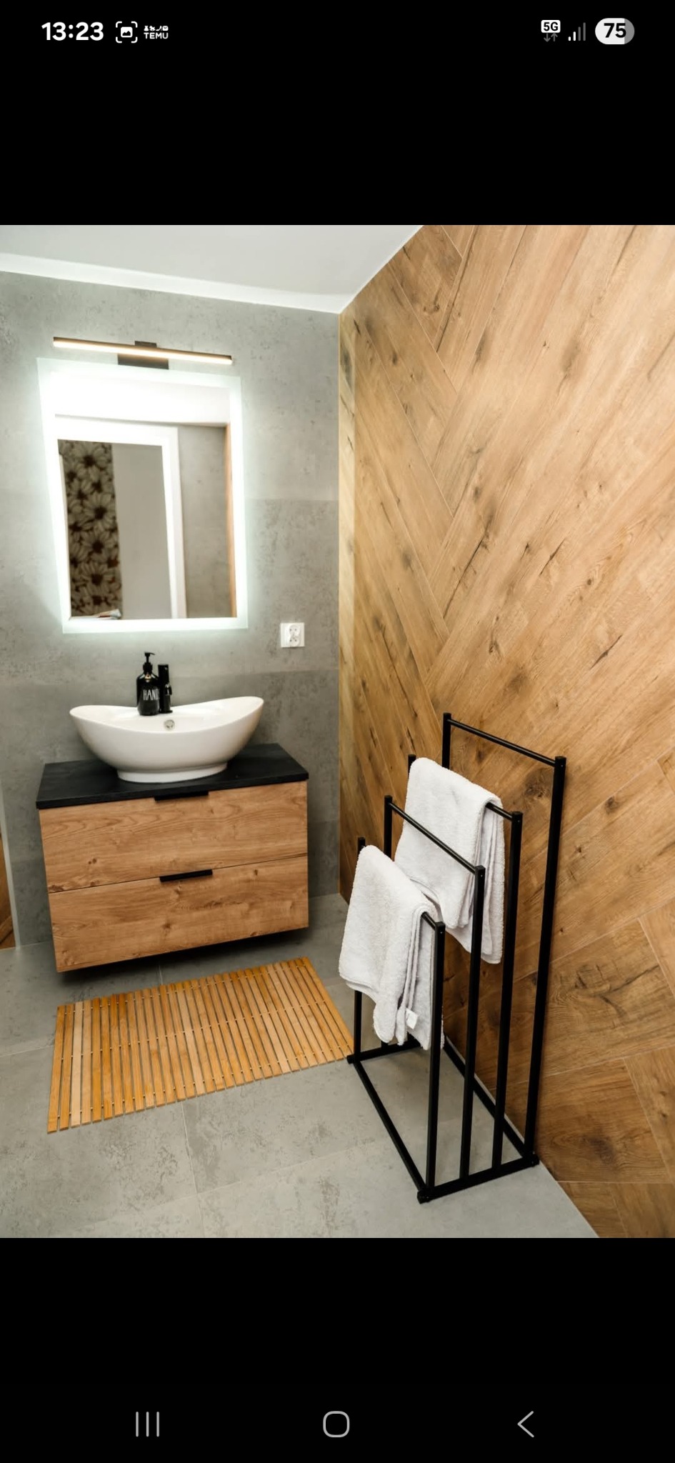 Nowoczesna łazienka z drewnianą ścianą w jodełkę, umywalką nablatową i podświetlanym lustrem. Na podłodze bambusowy dywanik, obok czarny wieszak na ręczniki.