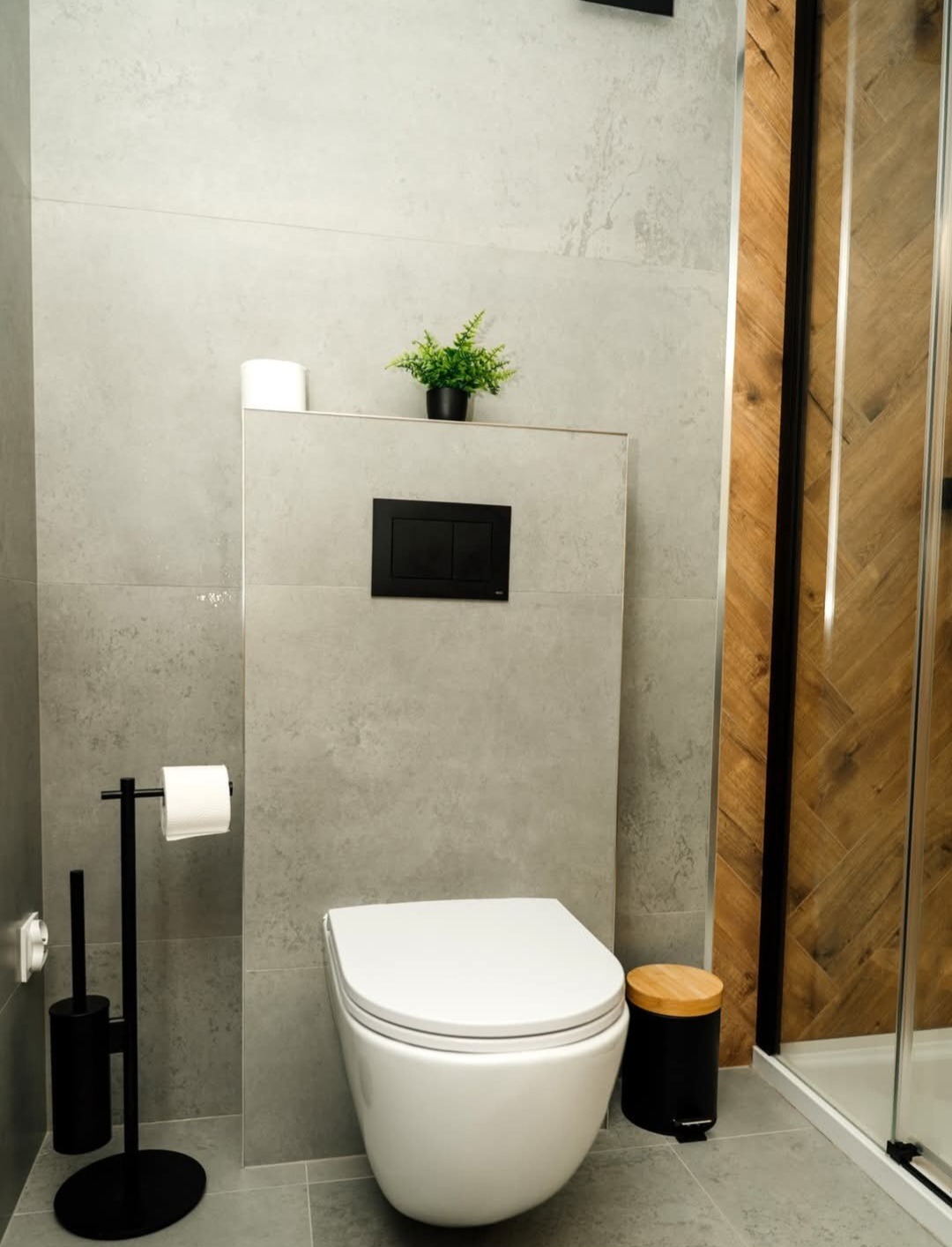 Nowoczesna łazienka z podwieszaną toaletą, szarą płytką i drewnianymi akcentami. Minimalistyczny design z rośliną doniczkową i czarnymi dodatkami.