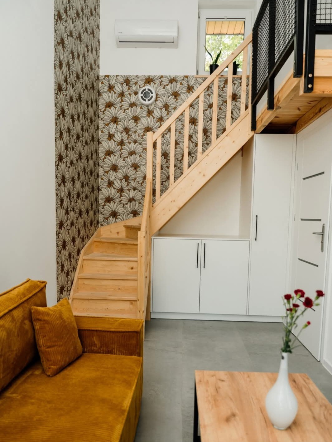 Jasne drewniane schody w nowoczesnym wnętrzu z wzorzystą tapetą i musztardową sofą. Minimalistyczny design, subtelne oświetlenie, kwiaty w wazonie na drewnianym stoliku.