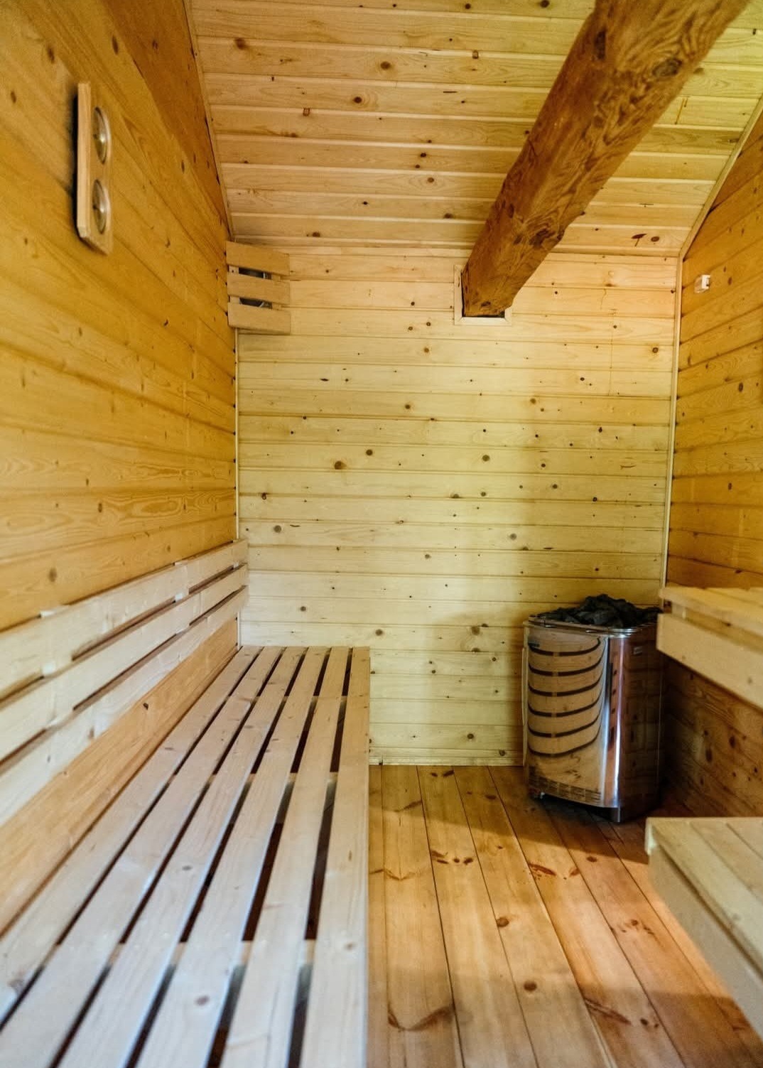 Drewniana sauna z ławkami, piecem i oświetleniem. Ściany i sufit wyłożone boazerią, widoczny fragment belki stropowej. Jasne, naturalne drewno.