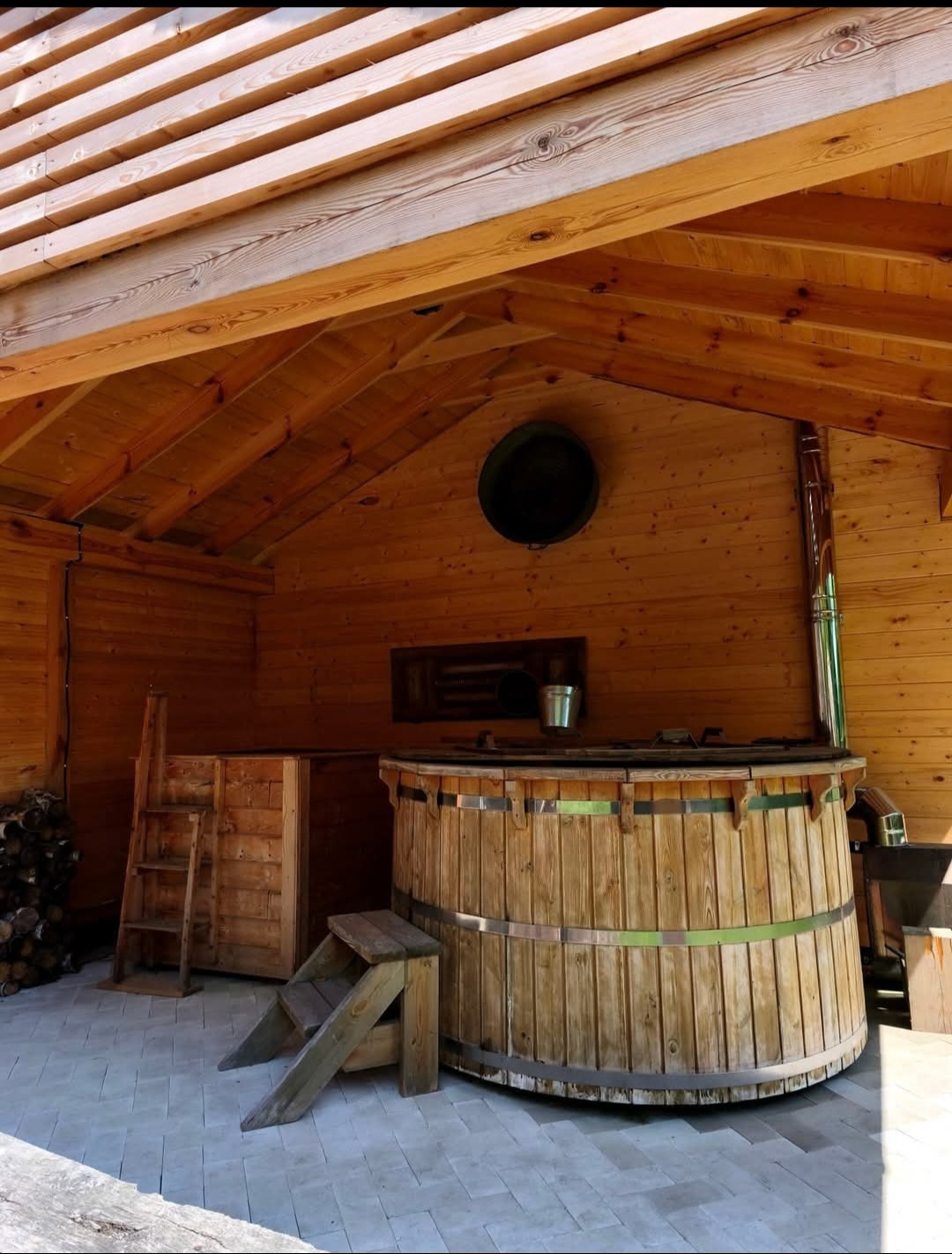 Drewniana balia z piecem wewnątrz drewnianej konstrukcji z dachem, widoczne schodki i komin. Całość utrzymana w ciepłej tonacji drewna.