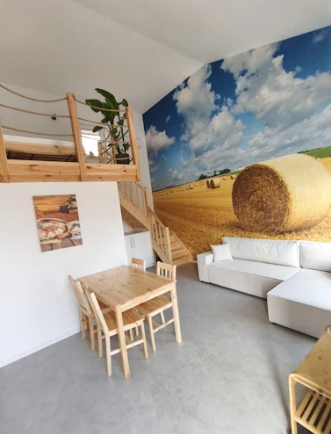 Jasne wnętrze z drewnianym stołem i krzesłami, białą sofą oraz fototapetą z motywem pola i nieba. Antresola z drewnianą barierką i schodami.