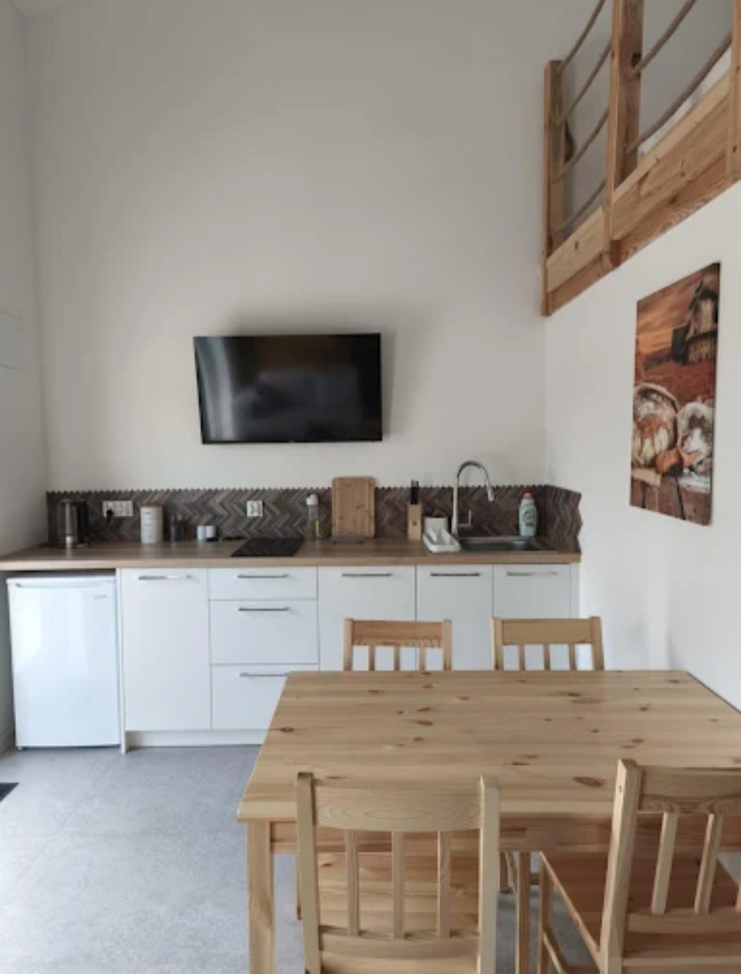 Jasna kuchnia z drewnianym stołem i krzesłami. Na ścianie telewizor i obraz. Widoczna antresola z drewnianą barierką. Minimalistyczny design.