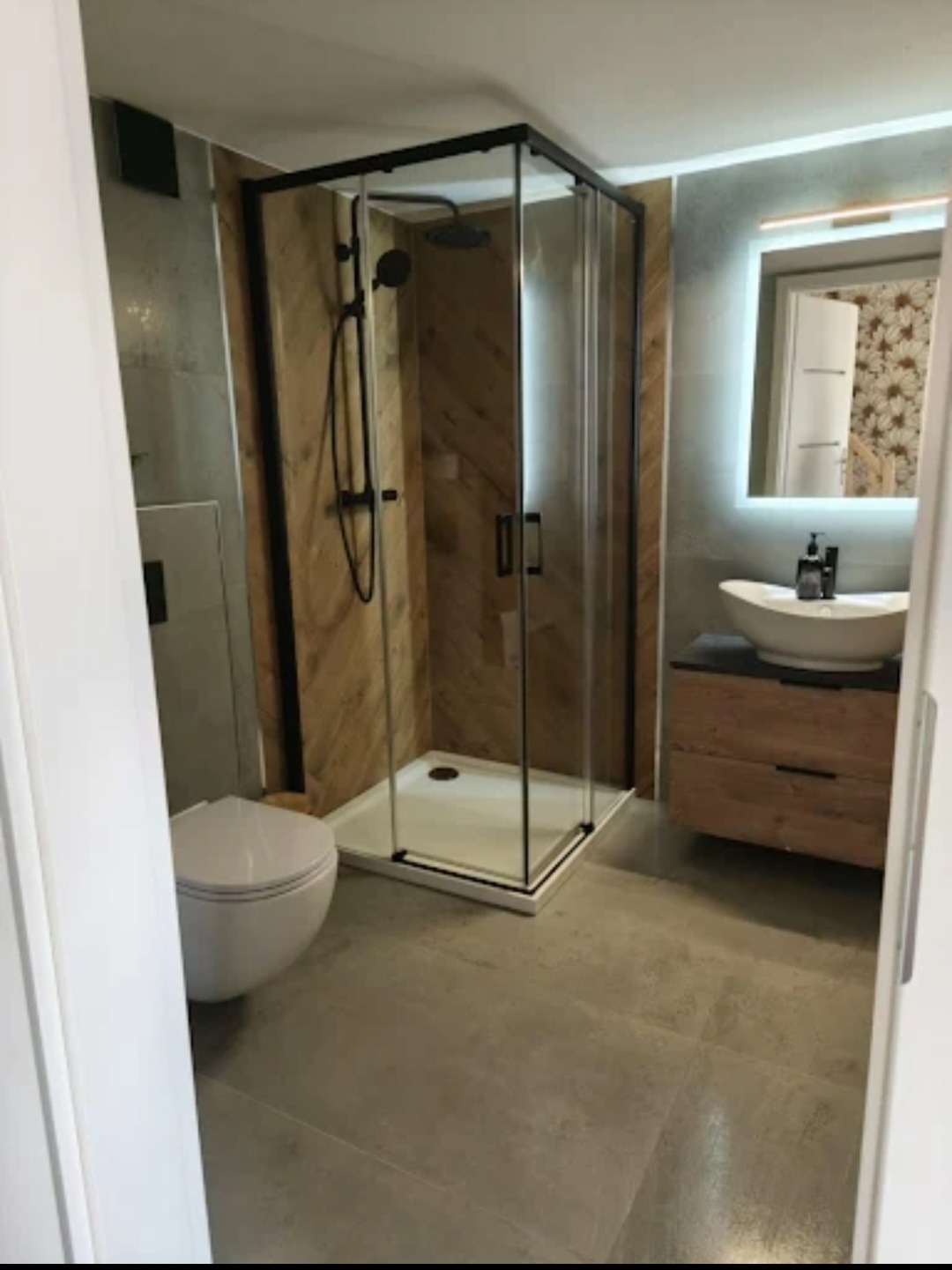 Nowoczesna łazienka z kabiną prysznicową typu walk-in z czarnymi profilami, umywalką na drewnianej szafce i podświetlanym lustrem. Widok zza uchylonych drzwi.