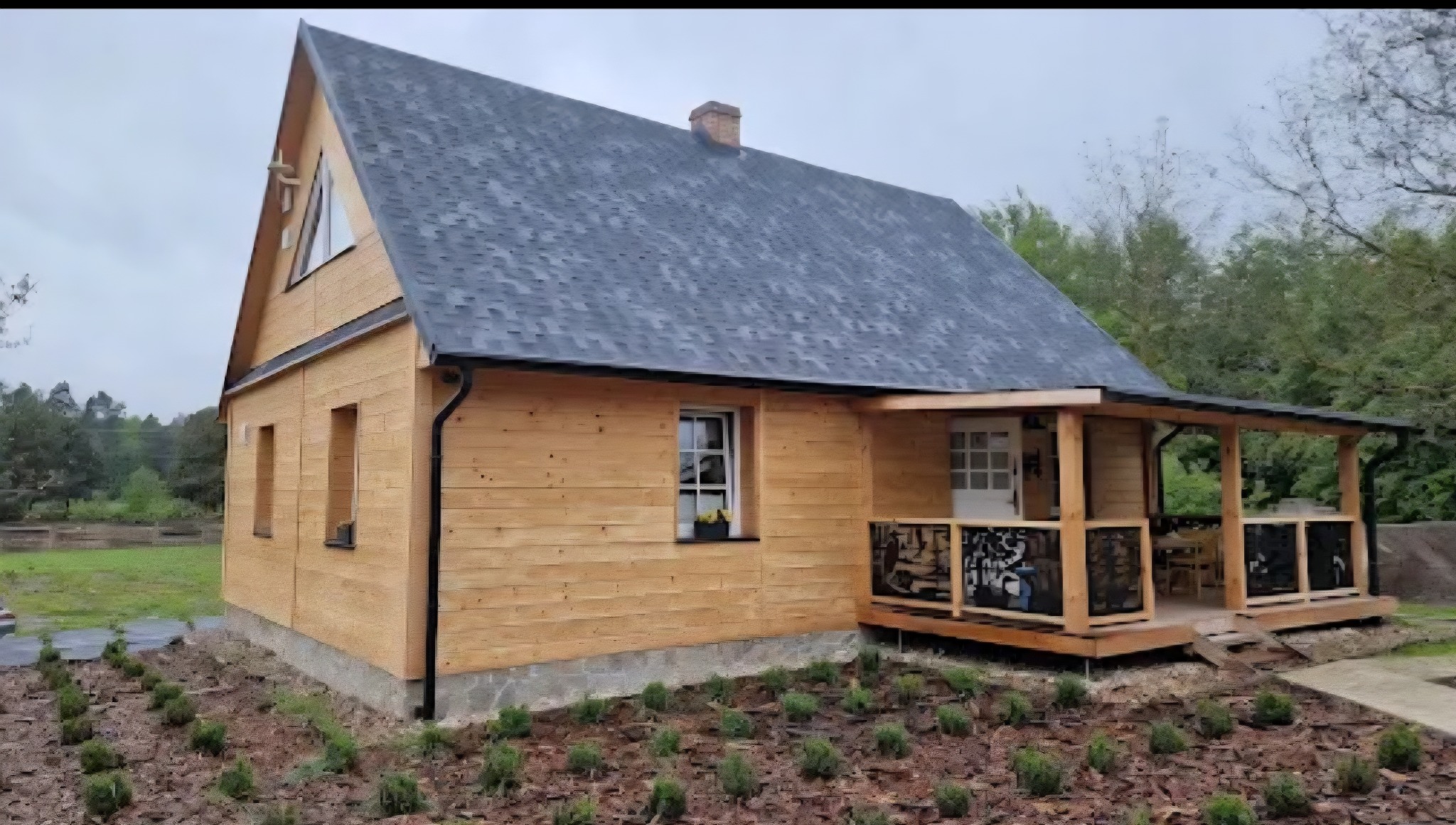 Drewniany dom z ciemnym dachem i werandą, otoczony zielenią. Widoczne detale wykończenia elewacji i dachu. Ujęcie z zewnątrz, w pochmurny dzień.