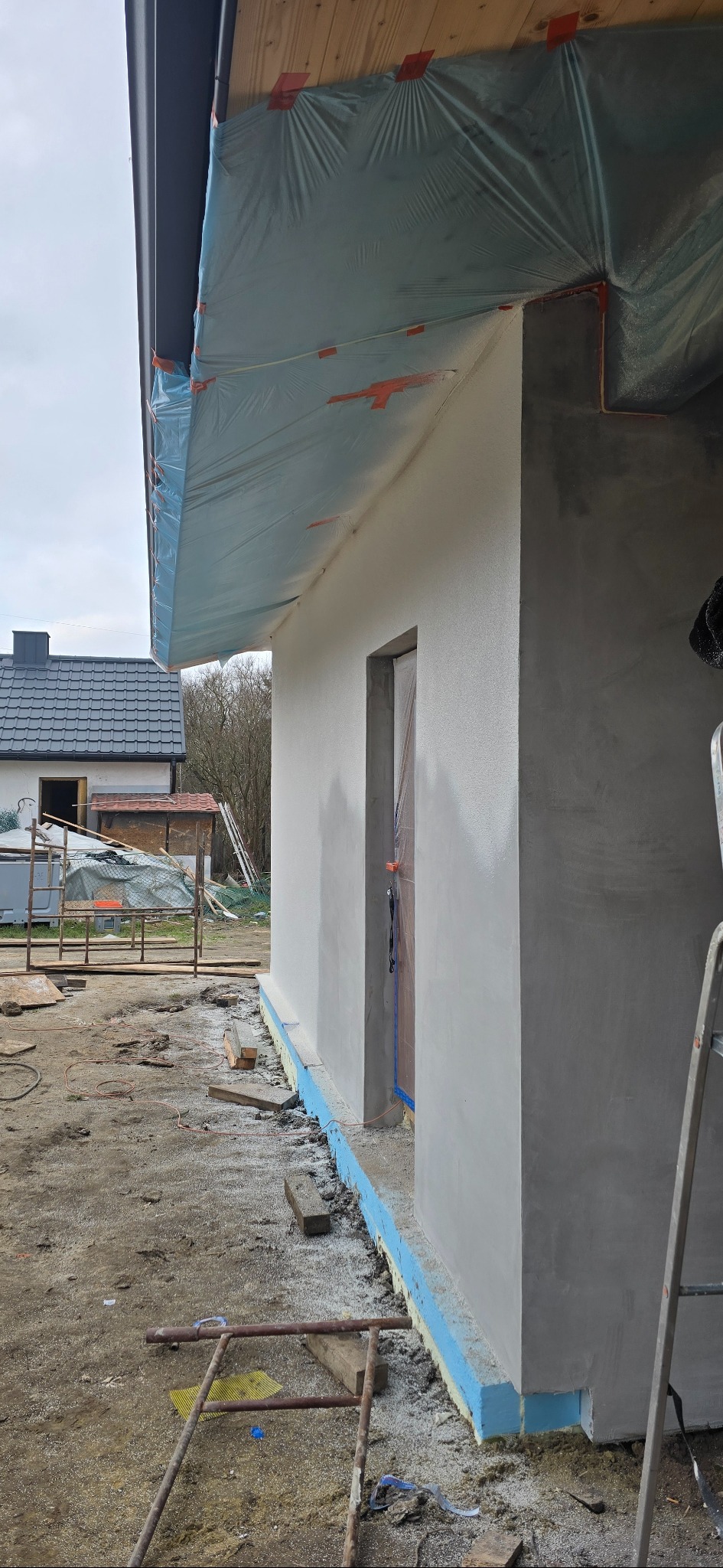 Ściana budynku po szpachlowaniu, z widocznym ociepleniem fundamentów i zabezpieczeniem folią dachu. Prace wykończeniowe w toku, drabina oparta o ścianę.