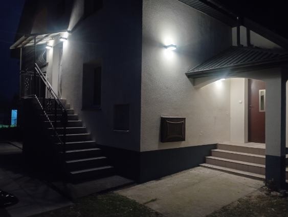 Oświetlony budynek z zewnętrznymi schodami i wejściem, nocne ujęcie z niskim kątem, widoczne detale elewacji i oświetlenia.