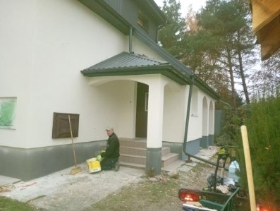 Pracownik z wiadrem farby klęczy przed wejściem do domu z pomalowaną elewacją. Widoczne narzędzia i fragmenty rusztowania. Świeżo pomalowana elewacja w jasnym odcieniu.