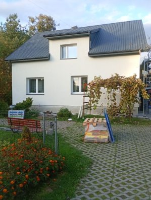 Biały dom z szarą blachodachówką po malowaniu elewacji, widok z ogrodu z kwiatami i ławką. Na ścianie pnącze, obok rusztowanie. Widoczne materiały budowlane.