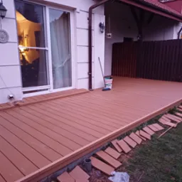 Nowo wybudowany taras z desek kompozytowych w kolorze jasnego drewna, częściowo ukończony, z widocznymi elementami montażowymi i narzędziami, przylegający do jasnego budynku z dużymi oknami.